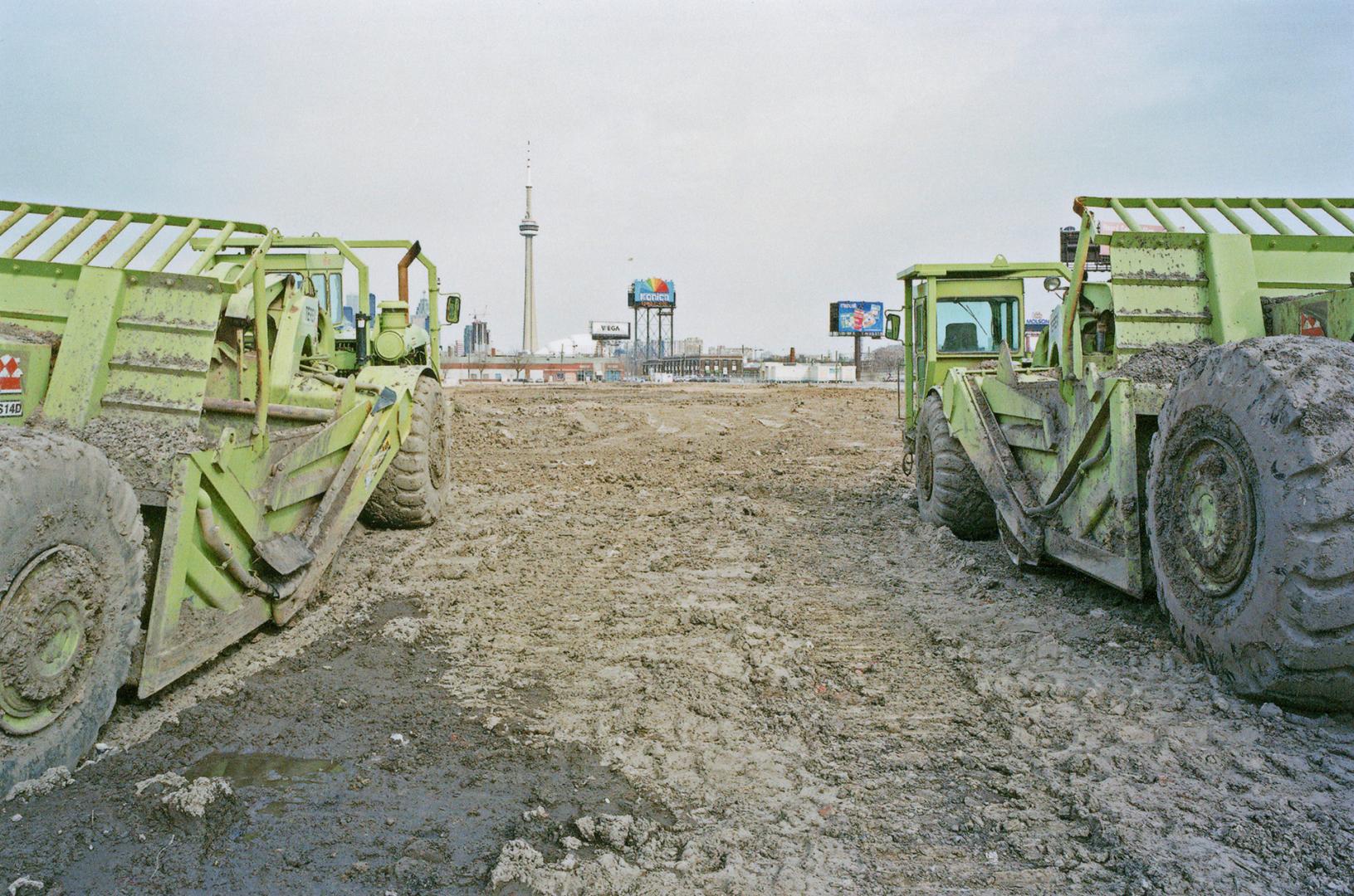 A photograph of two construction scrapers in a muddy field, with the CN Tower visible in the ba…