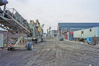 A photograph of a dirt laneway between buildings and construction trailers on the right side an…