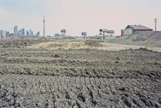 A photograph of a muddy field, with a shed visible to the right and the Toronto skyline includi…