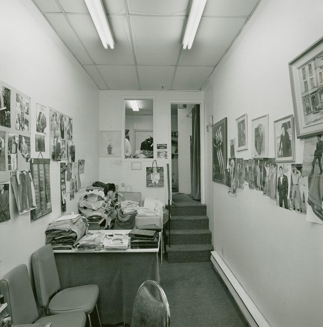 A photograph of a narrow room, with a desk, piles of clothing and fabric, mirrors and photograp…