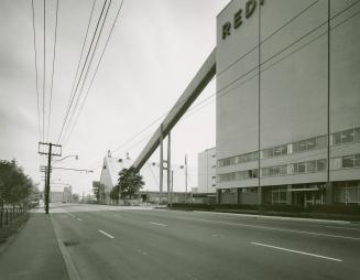 A photograph of a factory, with a chute leading from one building to another and a road in the …