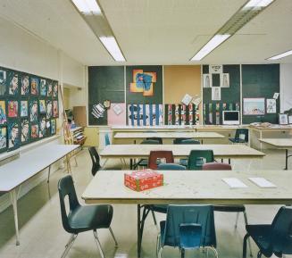 A view of a public school classroom, with tables, chairs and bulletin boards displaying artwork…