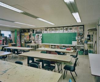 A photograph of a public school classroom, with chairs, tables, a bulletin board and artwork di…