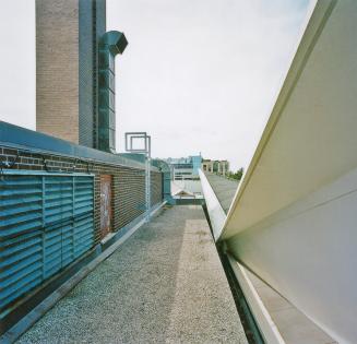 A photograph of a roof of a school, with air vents, a smokestack and apartment buildings in the…