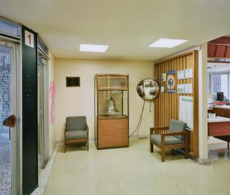 A photograph of an entrance foyer inside a public school, with a large bell inside a glass case…