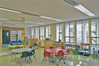 A photograph of a kindergarten classroom, with round tables and chairs, and windows facing out …