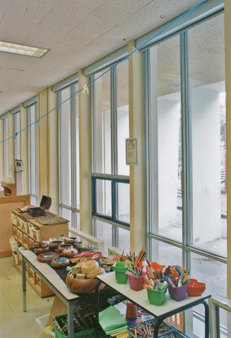 A photograph of a public school classroom, with tables and shelves in front of a wall of window…