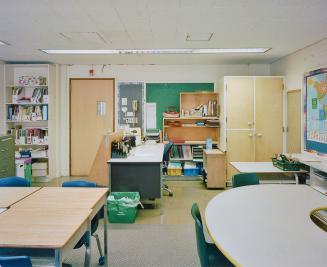 A photograph of a public school classroom, with chairs, tables, shelves and cabinets, a chalkbo…