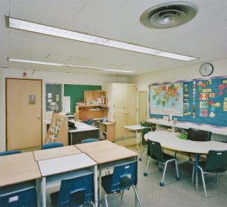 A photograph of a public school classroom, with chairs, tables, a chalkboard, shelves and cabin…