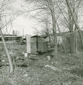 A photograph of a rudimentary wooden structure in a vacant lot, with buildings in the backgroun…