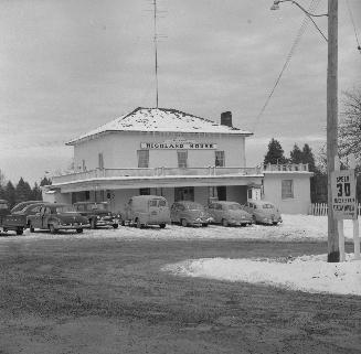 A photograph of a hotel with a sign reading &quot;HIGHLAND HOUSE,&quot; with cars parked in fro…