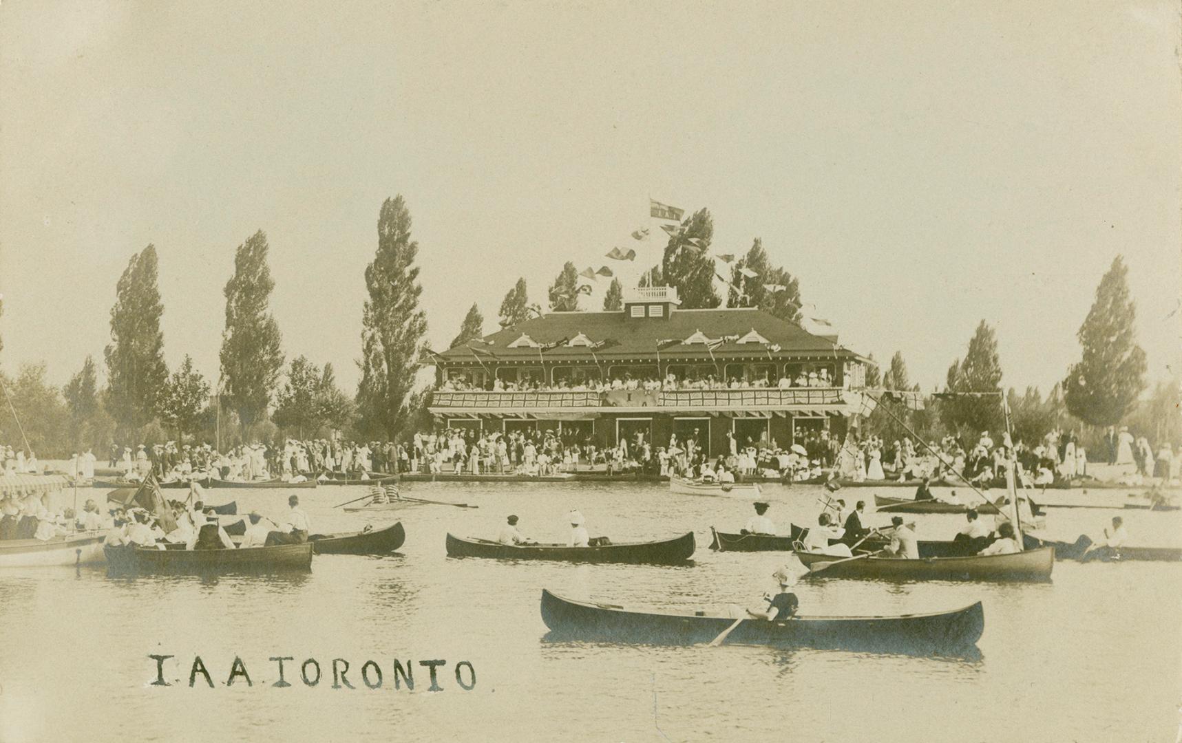 Black and white photographs of many canoes in the water in front of a three story Victorian bui ...