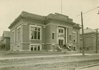 Black and white picture of public library building facing onto a dirt street with single lamppo…