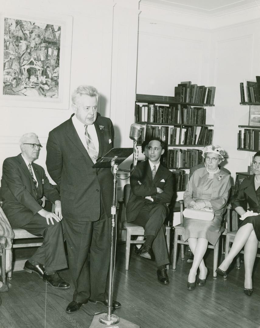 Sir Ernest MacMillan standing and speaking into a microphone with four other people sitting lis…