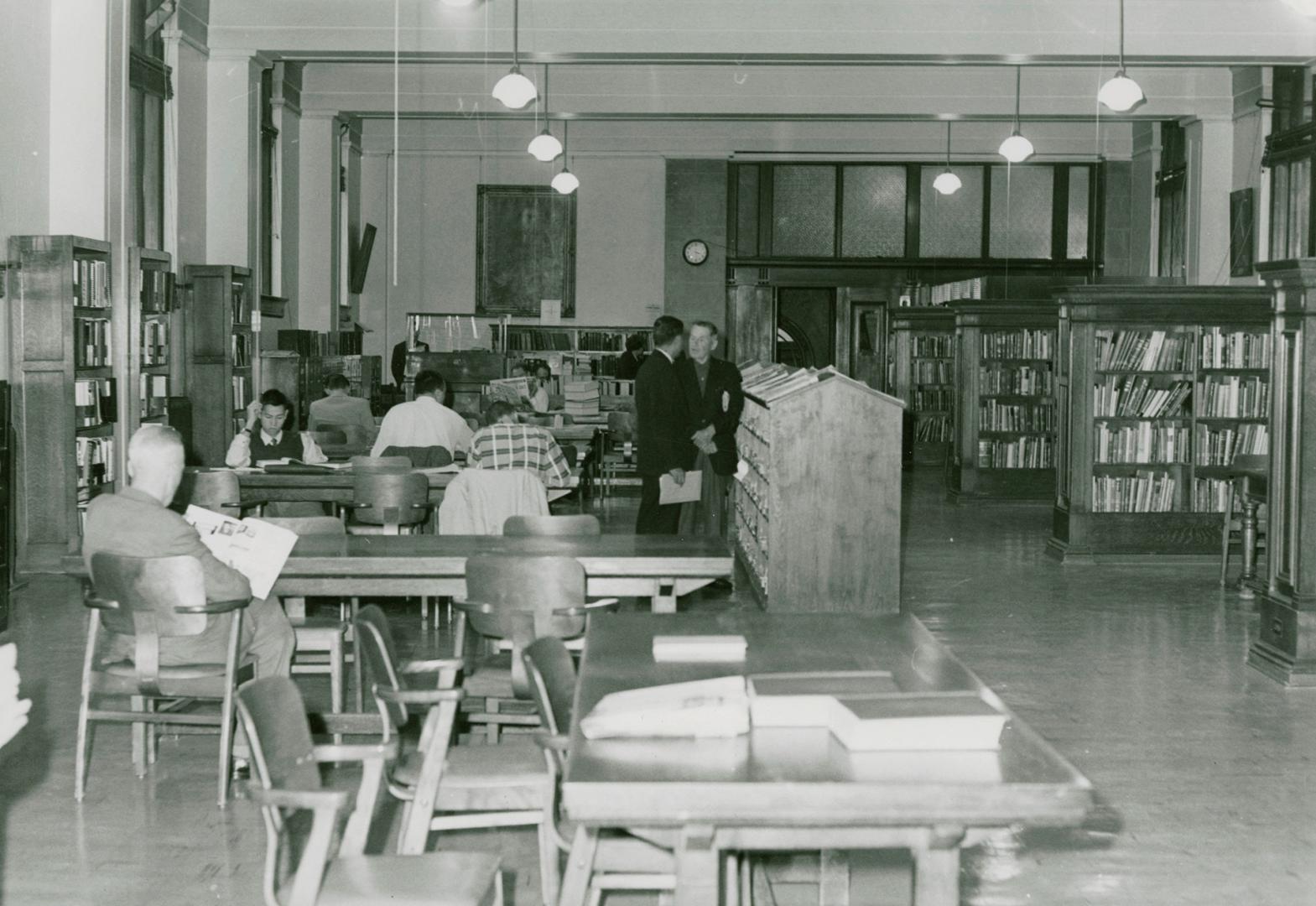 Picture of tables and chairs and people studying in a library room with rows of shelves of book…