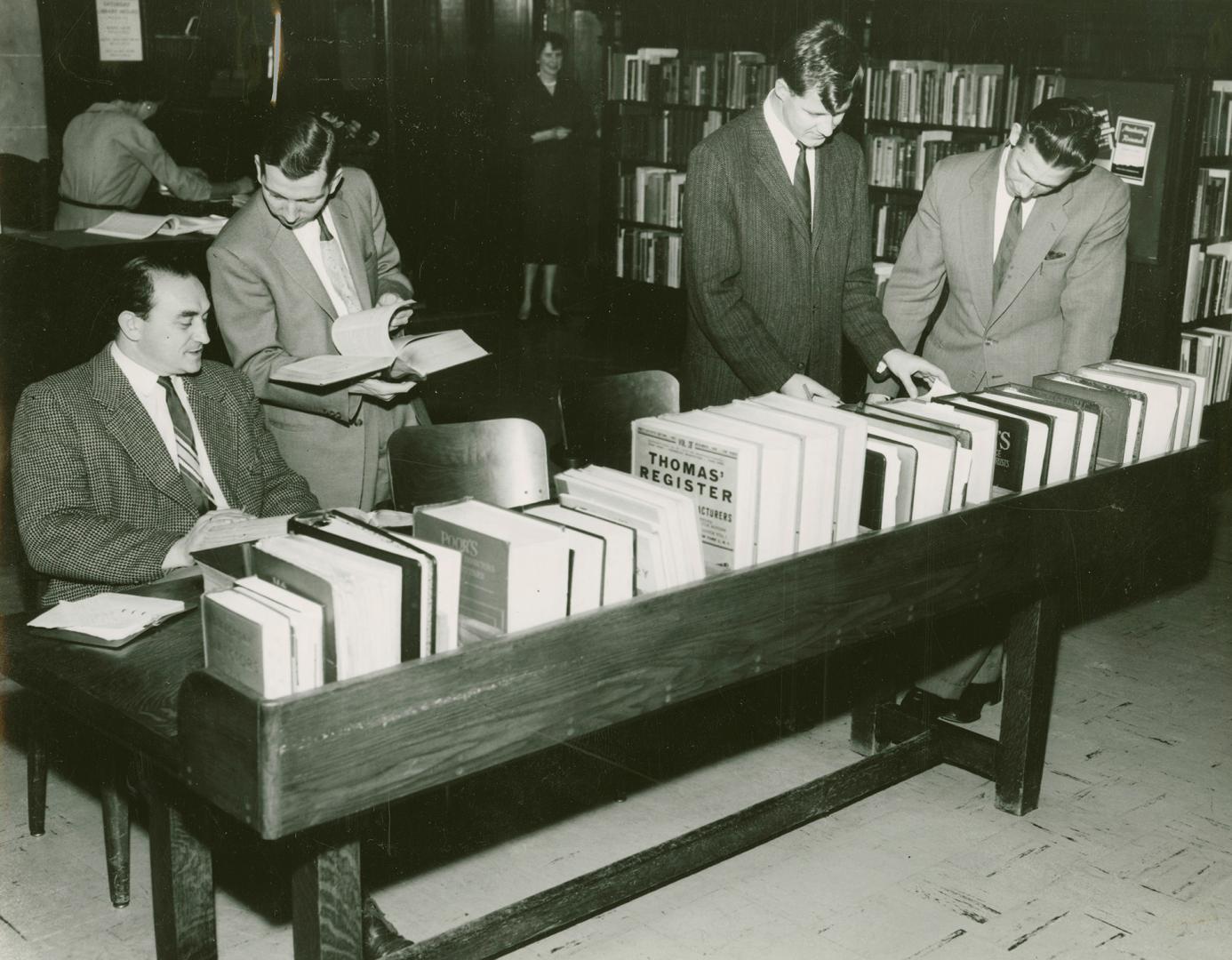 Three men in suits stand looking at a row of business directories and one man is consulting wit…