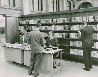 Two librarian, one male and one female, sit at a desk and assist a user while another user brow…