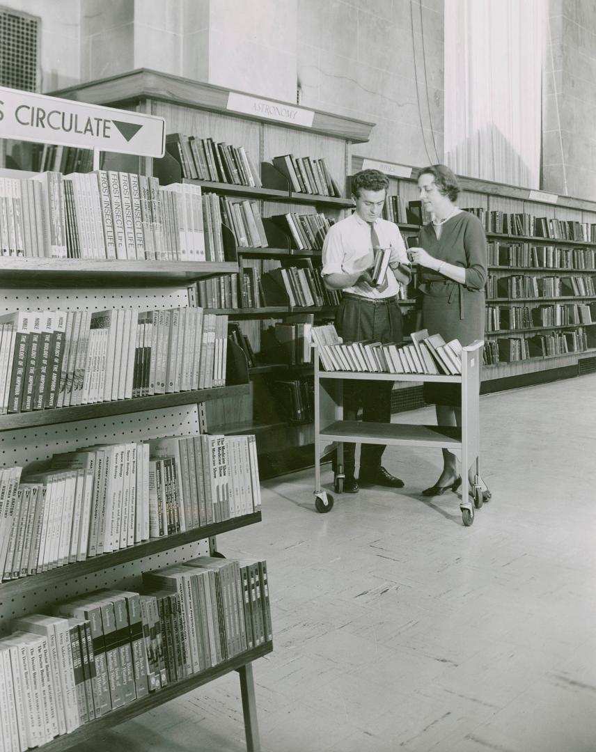 Two library staff, one man and one woman look at a book on a book truck while standing amongst …