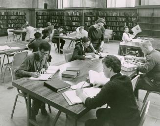 People sit at large tables studying in a large room lined with book shelves and the librarian s…