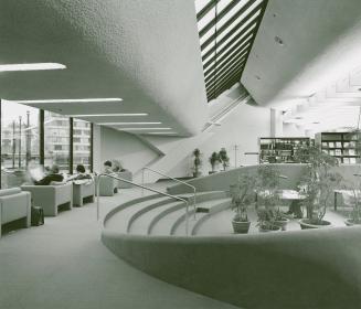 Interior of a library with sunken reading area containing plants and study tables and people si…