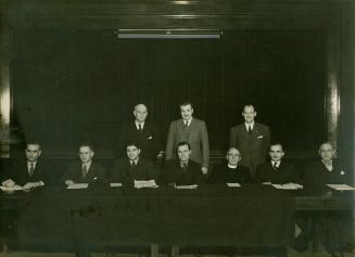 A posed photograph of ten men facing the camera, seven sitting at a long table and three standi…