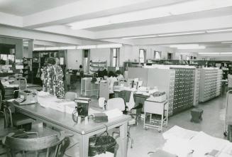 Picture of desks and card catalogues in a library work room with some staff in the background s…