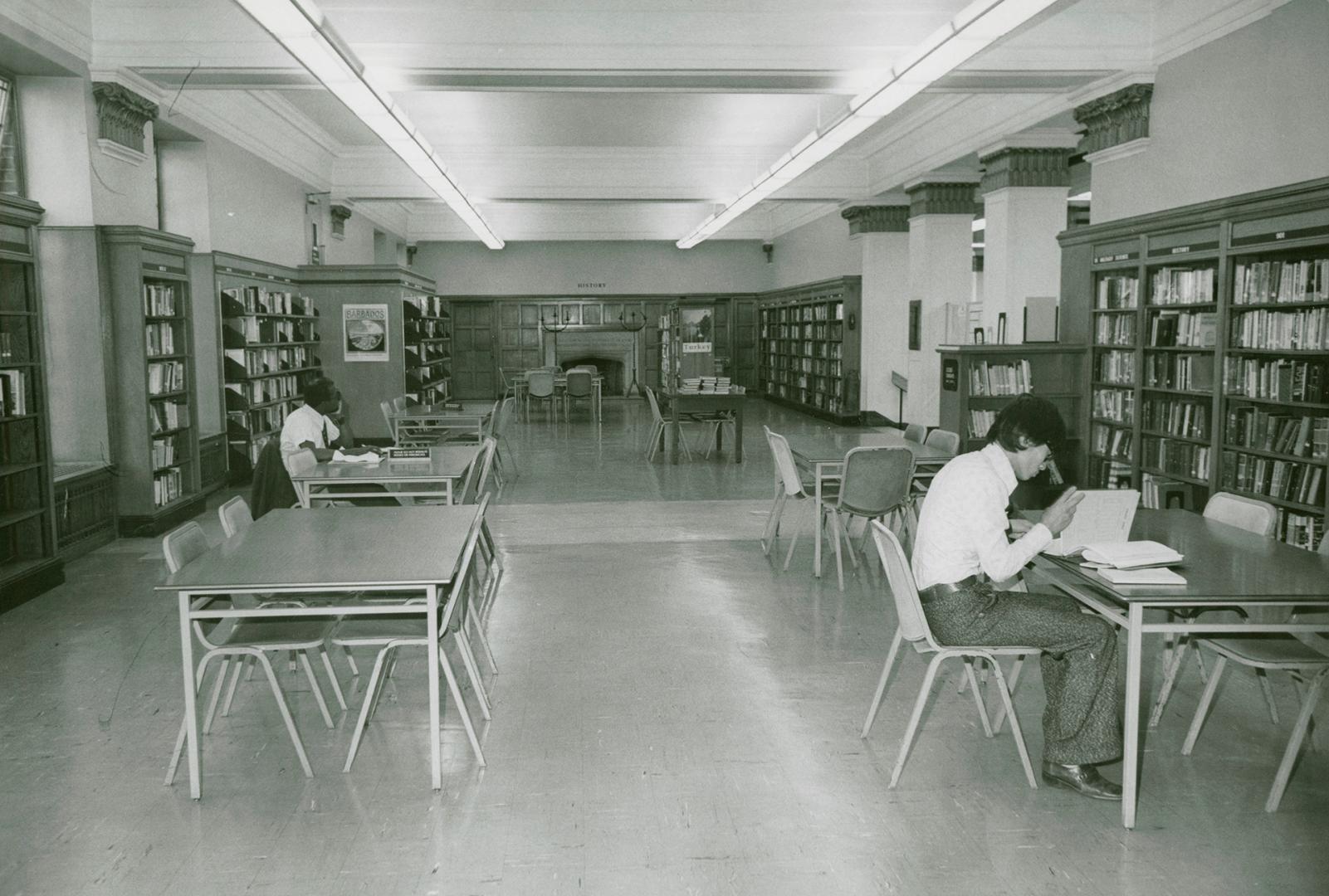 Library room with several tables and shelves of books around the walls with two people studying…