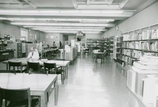 View of a library room with tables and shelves of books lining the wall and one man studying at…