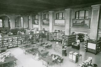 View of large library reading room with many tables, rows of shelves with books and large arche…