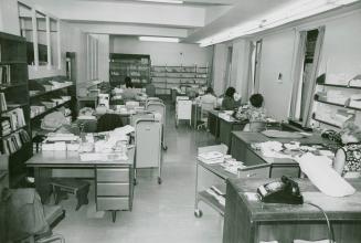 Library office work room with several desk and people seated working and several book trucks an…