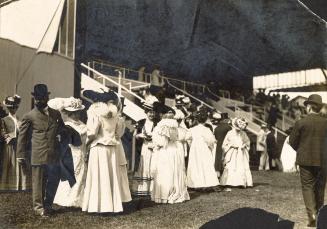 A group of people wearing formal clothing and hats stand on grass in front of the members' gran…