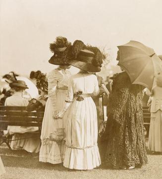 Three women wearing formal clothing and hats stand on grass in front of a bench that other simi…