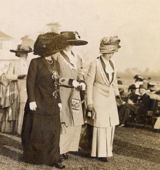 Three women wearing formal clothing and hats stand on grass in front of other, similarly dresse…