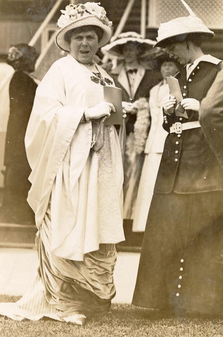 Two women wearing formal clothing and hats stand on grass front of a group of people who are dr…