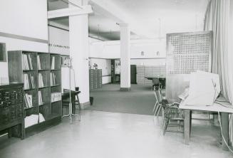 A sparsely furnished library room with tables with microfilm reading equipment, filing cabinets…