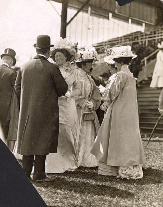 Four women and two men wearing formal clothing and hats stand on grass in front of a grandstand…