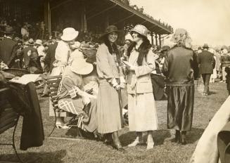 Two smiling women wearing formal dresses and hats stand in front of a large crowd of people at …