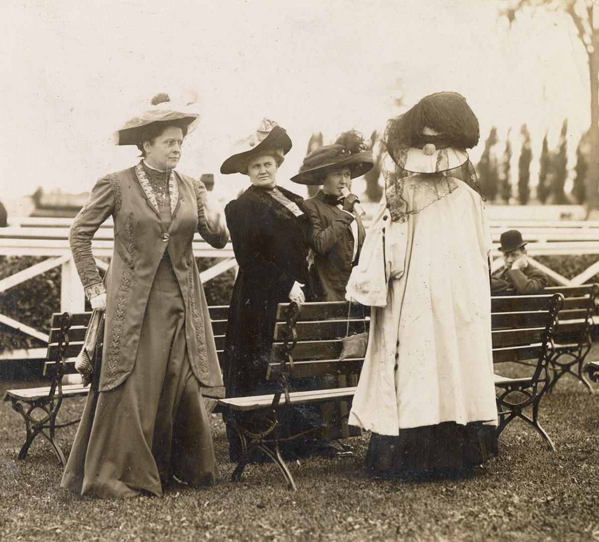 Four women wearing formal dresses and hats stand around two benches in front of a racetrack. On…