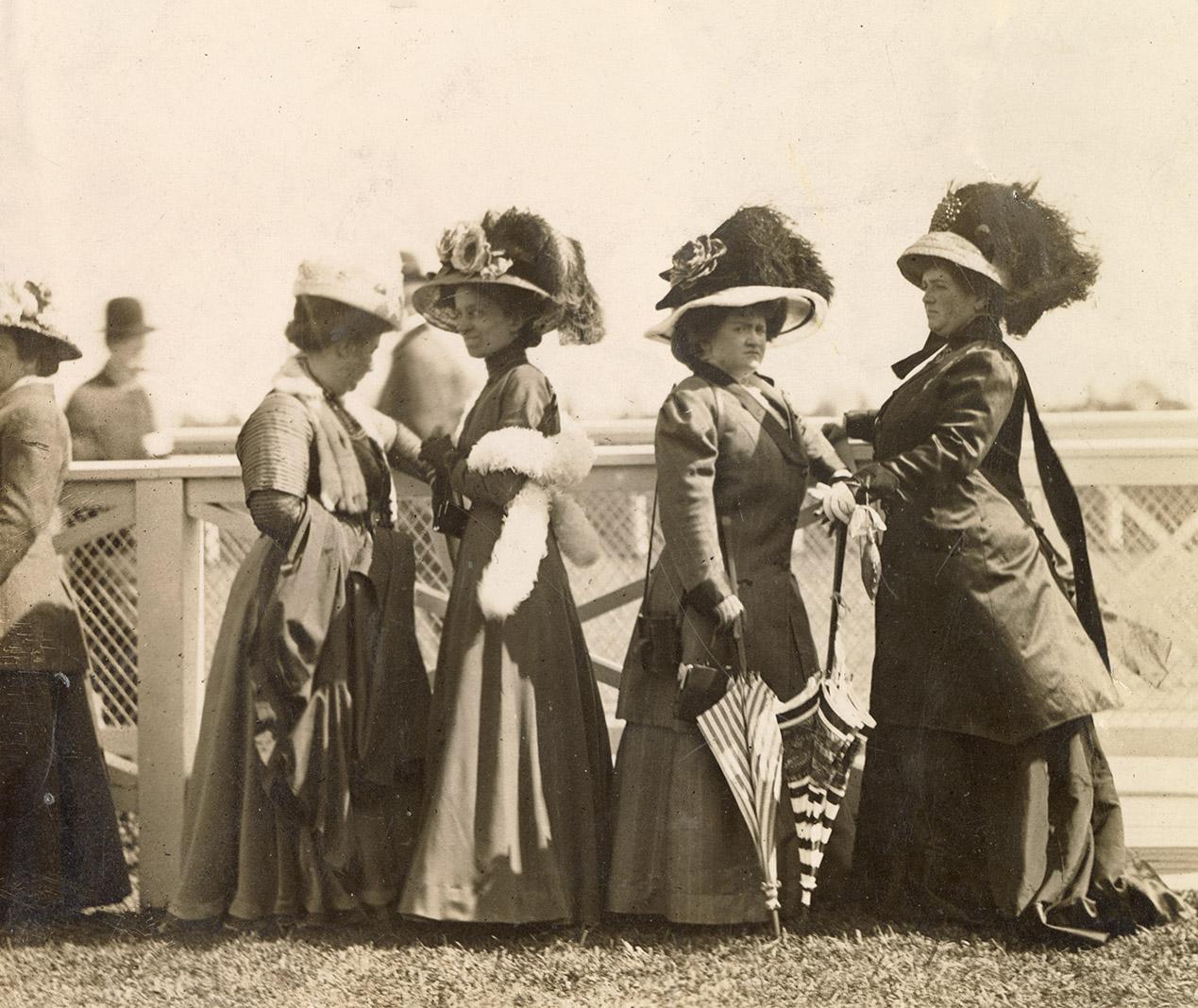 A group of five women wearing formal dresses and hats stand in front of a fence by a racetrack.…