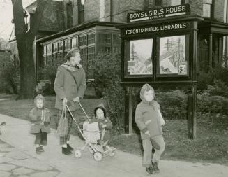 Photo of a woman and three children, one in a stroller, walking on the sidewalk past the Boys a…