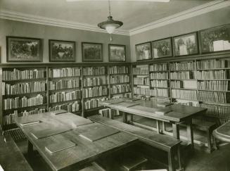 A room in Boys and Girls House lined with walls lined with books and two large wooden tables wi…