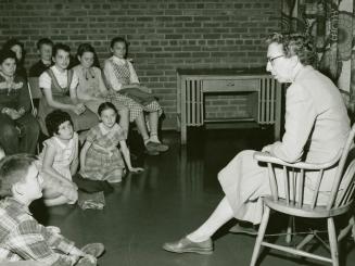 Picture of a librarian sitting in a chair reading telling a story to a group of children seated…