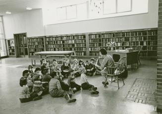 Photo of librarian reading a story to a group of children seated on the floor in a large room w…