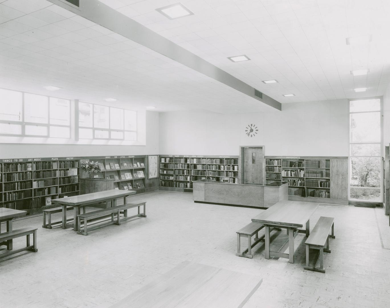 Photo of large room in a library with shelves of books lining the walls and tables and benches …