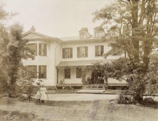 A cloudy picture of a two-storey house. A family group poses formally on chairs in the entryway…