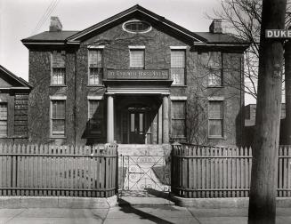 Photo of a stone house with nine sash windows and two storeys. There is a wooden picket fence a…