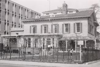 A 1970s photograph of a large, two and a half storey house with central chimney. A one-storey e…