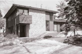 Photo of a modern two-storey brick house built on a hill, in a treed lot. Next to a driveway, t…