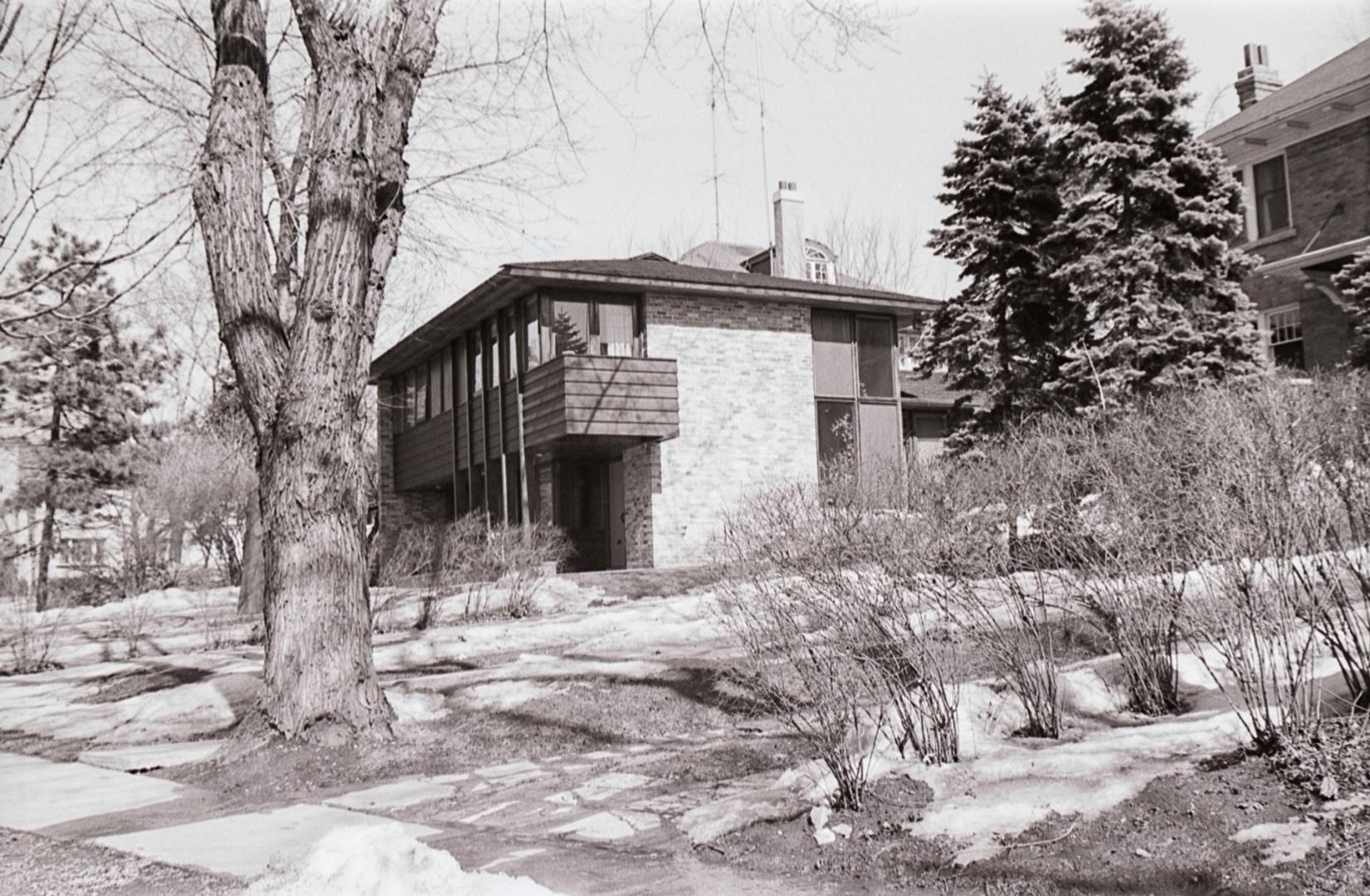 Photo of a modern two-storey brick house built on a hill, in a treed lot. Next to a driveway, t…