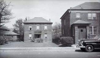 A photograph of a two-story residential house, with a road and a parked car in the foreground. …